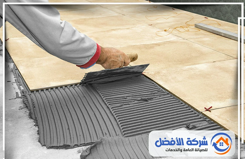 عامل يضع بلاطاً رخامياً بيج اللون على أرضية مغطاة بمونة لاصقة، ويستخدم ميزان التسوية.