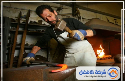حدادة فنية بجدة حداد يرتدي مريلة وقفازات، يستخدم مطرقة لتشكيل قطعة معدنية ساخنة على السندان.