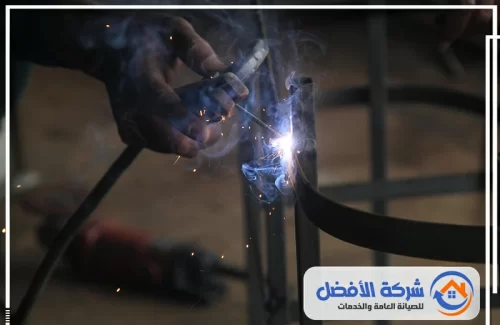 ورشة حدادة أبواب وشبابيك المدينة المنورة لقطة مقربة ليد حداد يلحم قطعة معدنية، مع توهج شديد للشرر.