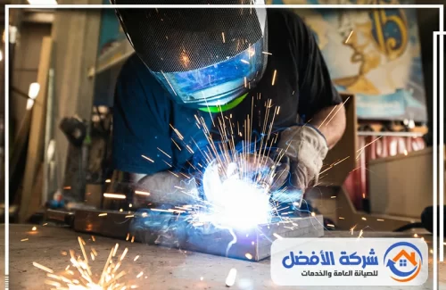 تفصيل أبواب حديد المدينة المنورة حداد يرتدي خوذة واقية أثناء لحام معدن، مع تصاعد الشرر والدخان.