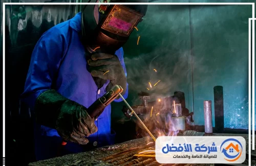 حداد المدينة المنورة عامل يرتدي قناع لحام وهو يقوم بتشكيل المعدن عبر عملية اللحام، وسط توهج قوي.