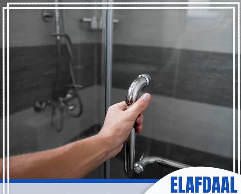 تركيب شاور بوكس سيكوريت في جدة 0547648005 شركة الأفضل يدًا تمسك بمقبض باب كابينة شاور زجاجية داخل حمام، مع ظهور الدش والزجاج الشفاف في الخلفية.