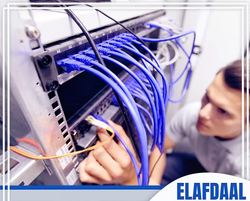 فني شبكات يعمل على توصيل وتنظيم كابلات، مع وجود منافذ شبكة مستخدمة في تمديدات الإنترنت والبيانات.