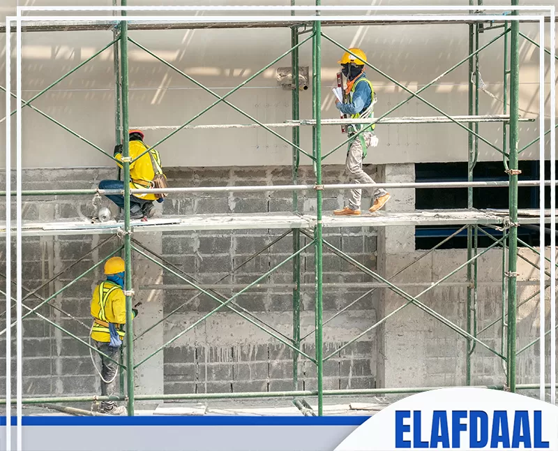 عمال تشطيبات يعملون على سقالات خارجية، مع ارتداء خوذات وسترات أمان أثناء تنفيذ أعمال دهان أو معالجة واجهة مبنى قيد التشطيب.